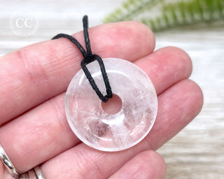 A hand holding a clear Quartz donut pendant on a black cord 