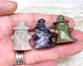 A hand holding three crystal Ghost with Witches Hat carvings in various crystal types 