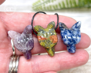 A hand holding three wolf head carvings, from left to right - one Amethyst, one Unakite and one Sodalite. The Unakite Wolf head in the middle is on a black cord.